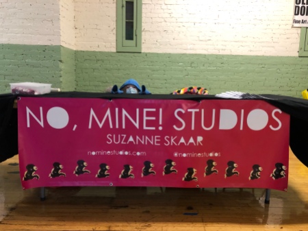 Picture of child in hoodie hiding behind table. On the front of the table is a large banner which reads "No, Mine! Studios; Suzanne Skaar; nominestudios.com; @nominestudios". A row of pixel art dinosaurs runs along bottom of banner.